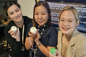 Three people smiling and holding branded cupcakes in front of a screen displaying business news in a casual office setting.