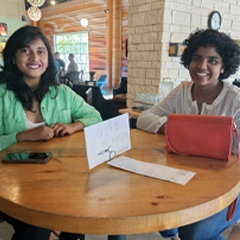 Two people sitting at a wooden table in a cozy restaurant, smiling warmly at the camera with a sign in front of them.