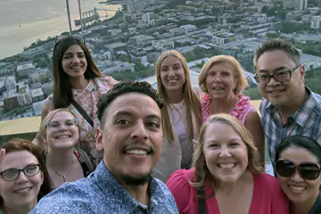 Ein Gruppen-Selfie von lächelnden Mitarbeitenden mit einer Stadtlandschaft im Hintergrund, aufgenommen von einem hohen Aussichtspunkt bei Sonnenuntergang.