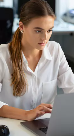 Junge Person in einer weißen Bluse, die an einem Schreibtisch am Laptop arbeitet.