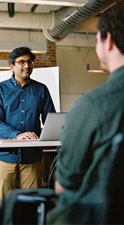Person wearing glasses standing at a desk with a laptop, speaking to a seated colleague.