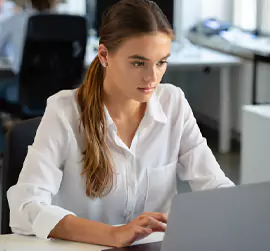 Young person in a white blouse working on a laptop at a desk.
