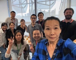 Group selfie of coworkers gathered around a table with food, smiling and flashing peace signs.